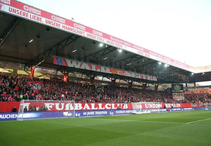 Trybuna na stadionie Union Berlin wypełniona kibicami. Na czerwonym banerze widnieje napis "UNSERE LIEBE. UNSERE MANNSCHAFT. UNSER STOLZ. UNSER VEREIN.".