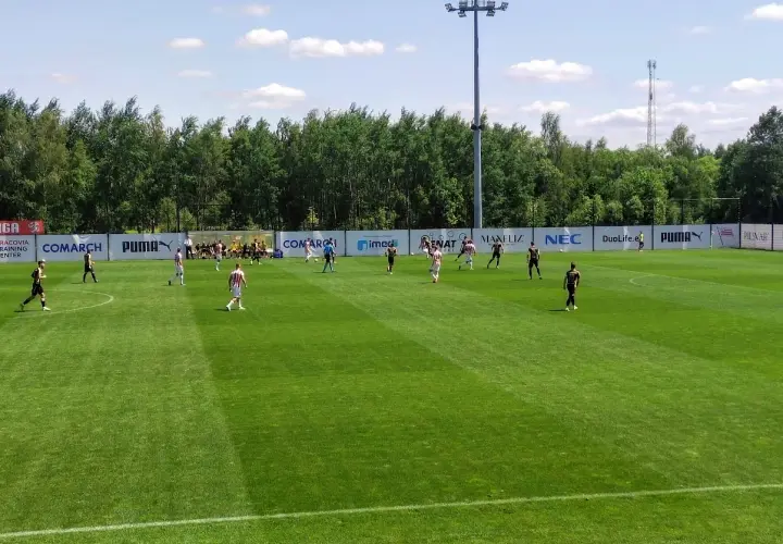 Piłkarze grają na boisku podczas meczu Betclic 3 liga. Słońce świeci, a drzewa tworzą tło.