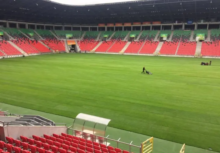 Pracownicy koszą trawę na boisku stadionu GKS Tychy. Puste trybuny w kolorach czerwonym i zielonym czekają na kibiców.