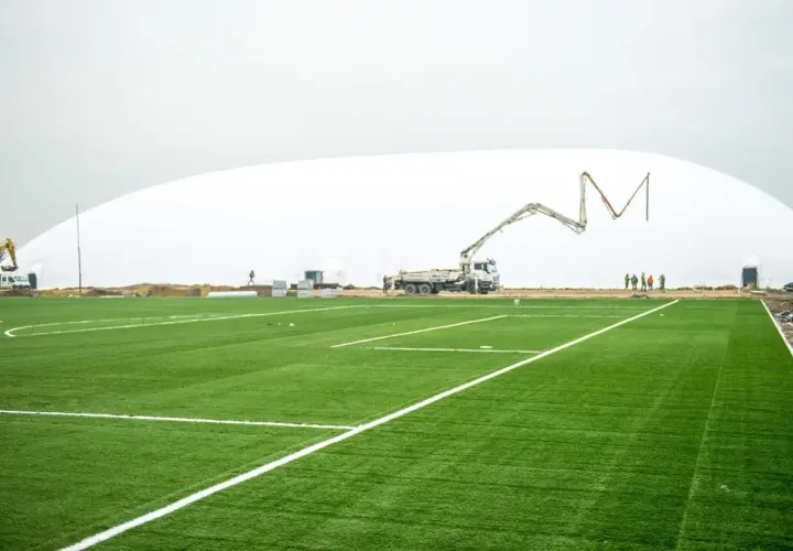 Budowa hali pneumatycznej na stadionie Ślęzy Wrocław. Sztuczna murawa z białymi liniami, koparka i gruszka do betonu w tle.
