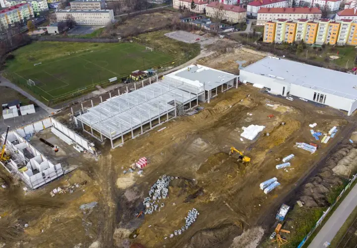 Budowa obiektu sportowego w Radzionkowie. Widać plac budowy z fundamentami, konstrukcją hali, maszynami budowlanymi i pobliski stadion.