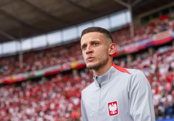 Sebastian Szymański w szarej bluzie z orłem na piersi, na tle trybun wypełnionych kibicami w biało-czerwonych barwach.