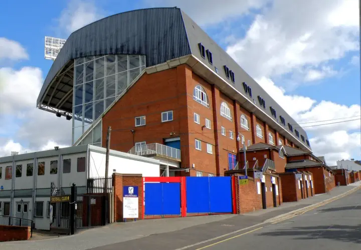 Widok na ceglany budynek stadionu Crystal Palace z charakterystycznym zadaszeniem i przeszkloną ścianą.