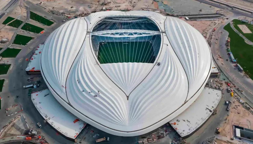 Al Janoub Stadium z lotu ptaka, z charakterystycznym białym dachem przypominającym żagiel. Widać zieloną murawę boiska i otaczające go tereny budowy.