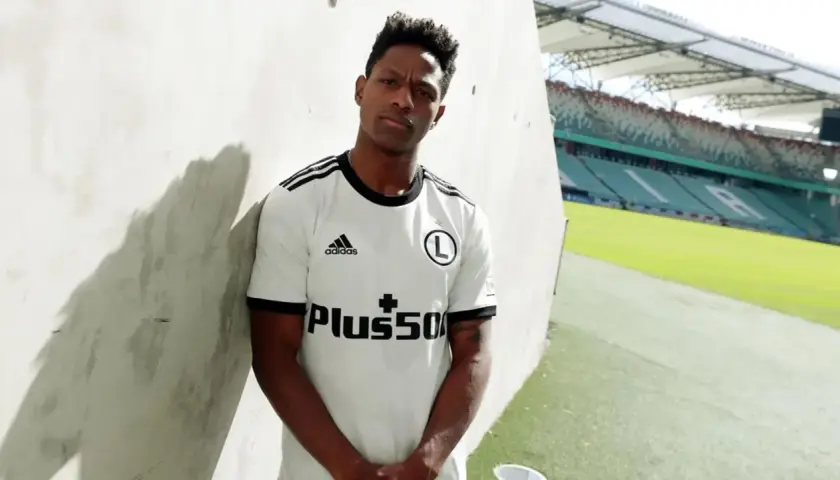 Joel Valencia w białej koszulce Legii Warszawa, z logo Plus500, na tle stadionu.