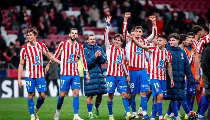 Piłkarze Atletico Madryt w pasiatych koszulkach świętują zwycięstwo. Wszyscy uśmiechnięci, niektórzy unoszą ręce w geście radości, kiedy gra Atletico Madryt przynosi sukces.