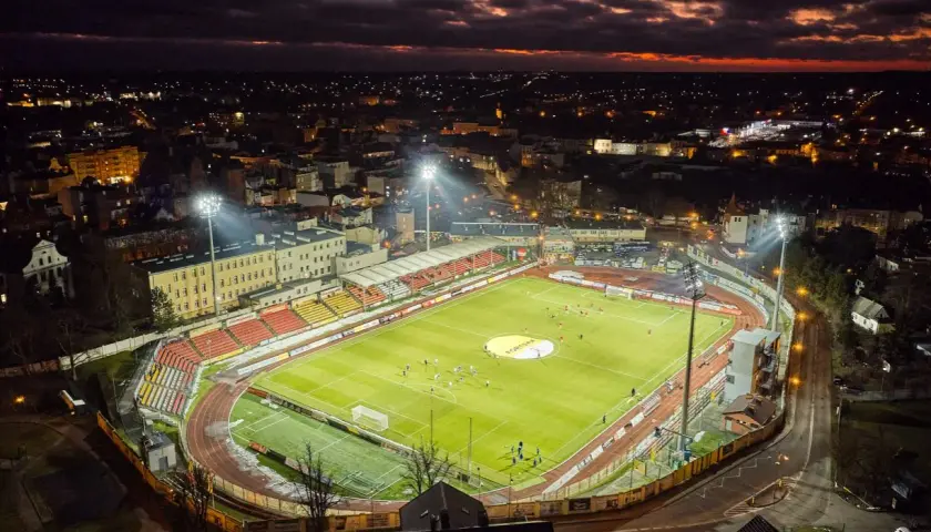 Wieczorny mecz na stadionie miejskim w Chojnicach. Piłkarze biegają po jasno oświetlonej murawie, a nad miastem zapada zmrok.