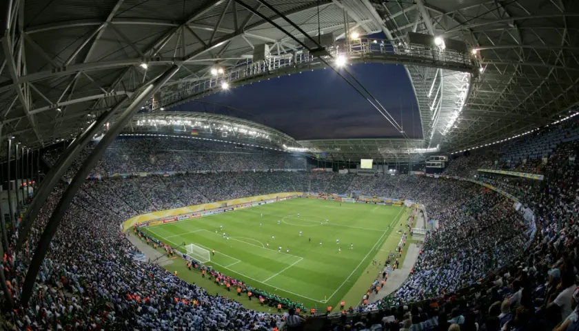 Widok z góry na pełny stadion Red Bull Arena Leipzig, gdzie tysiące kibiców w zielonych i niebieskich barwach dopingują swoje drużyny podczas nocnego meczu.