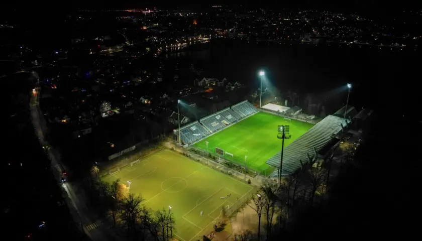 Nocne oświetlenie rozświetla zieloną murawę stadionu Sokoła w Ostródzie, otoczonego ciemnością i miejskimi światłami.