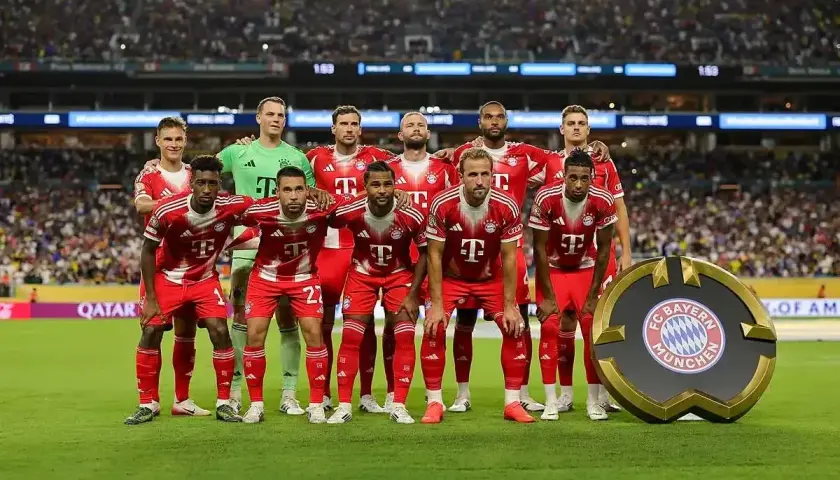 Drużyna Bayern Monachium – zawodnicy w czerwonych strojach pozują do zdjęcia na tle trybun. Obok nich stoi okrągłe logo klubu.