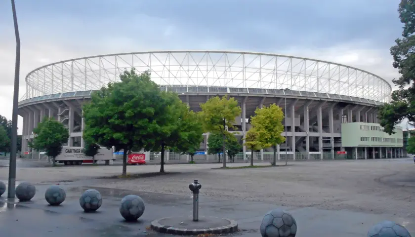Ernst Happel Stadion z charakterystyczną białą kratownicą dachu, otoczony zielenią i betonowymi kulami przypominającymi piłki nożne.