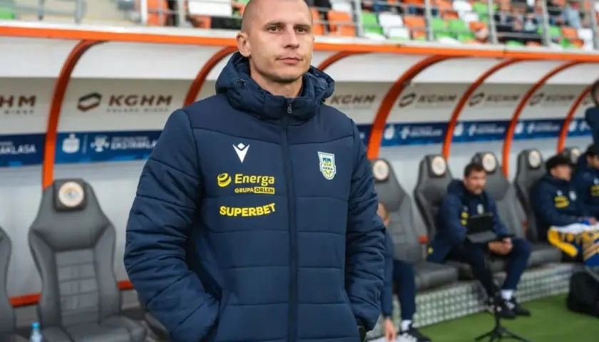 Trener Arki Gdynia, w kurtce z logo Energa i Superbet, stoi na tle ławki rezerwowych.