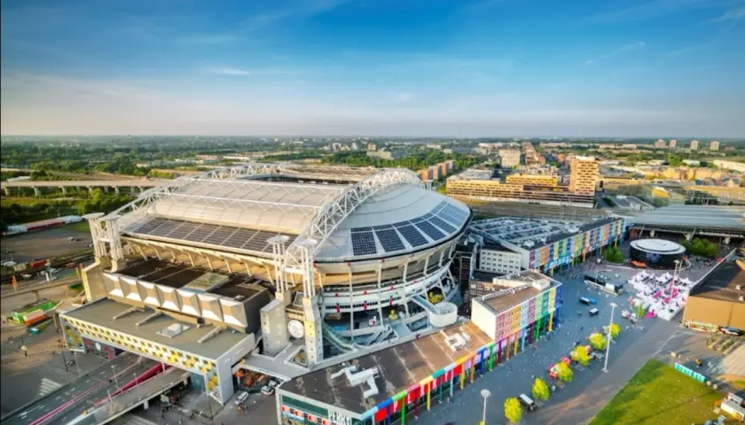 Johan Cruyff Arena, imponujący stadion z panelami słonecznymi na dachu, otoczony kolorowymi budynkami i zielenią.