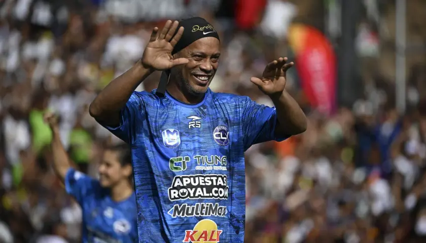 Uśmiechnięty Ronaldinho w niebieskiej koszulce z logo "FUTBOL CL" macha do kibiców. Widać, gdzie gra Ronaldinho.