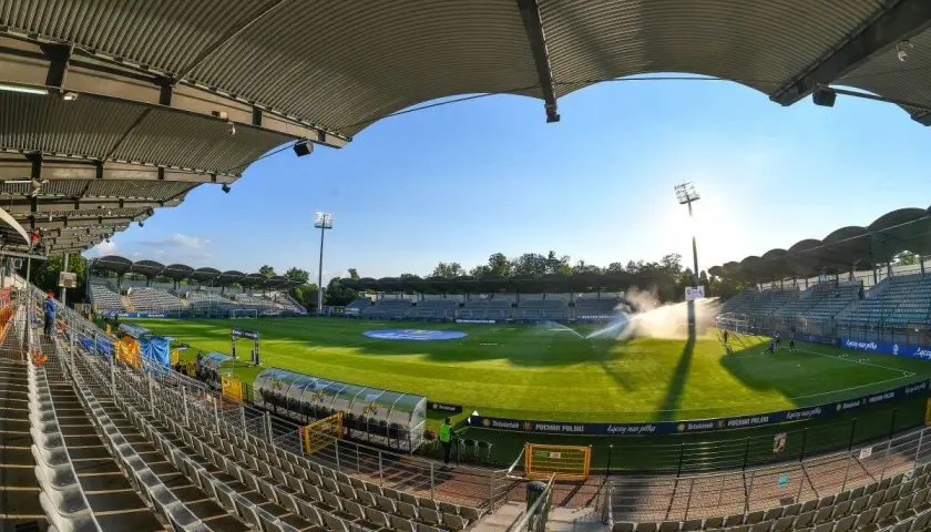 Pusta trybuna stadionu Miedzi Legnica, zielona murawa i zraszacze tworzą spokojny obraz przed meczem.