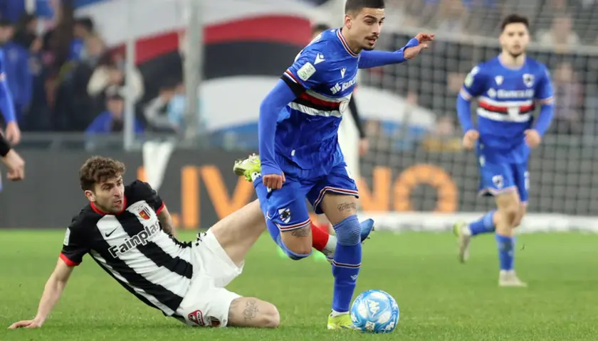 Piłkarze w akcji podczas rozgrywek Serie B. Jeden z zawodników w niebieskim stroju próbuje odebrać piłkę leżącemu na trawie przeciwnikowi w czarno-białej koszulce.