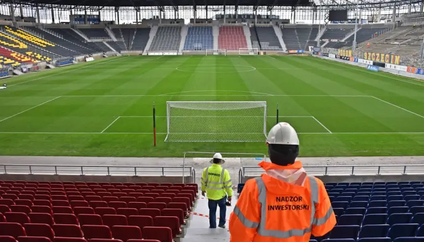 Pusta trybuna stadionu Pogoni Szczecin, z dwoma pracownikami w kamizelkach odblaskowych.