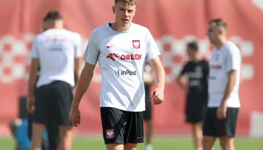 Szymon Żurkowski w białej koszulce reprezentacji Polski z logo Orlen i InPost, na tle czerwonej bandy.