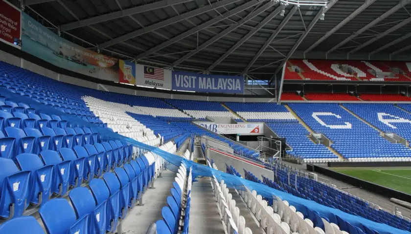 Puste trybuny Cardiff City Stadium, z niebieskimi i białymi siedzeniami, ozdobione banerem "Visit Malaysia".