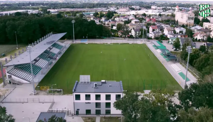 Stadion Wisłoki Dębica z zieloną murawą i trybunami, otoczony drzewami i zabudowaniami miejskimi.