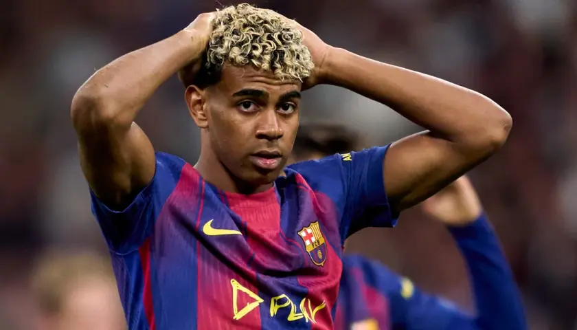Młody piłkarz Lamine Yamal w koszulce FC Barcelona z blond lokami, trzymający się za głowę.