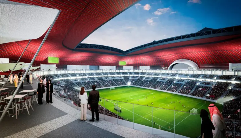 Widok na zieloną murawę stadionu Al Bayt, otoczoną trybunami pełnymi widzów. Czerwony dach tworzy imponującą konstrukcję.