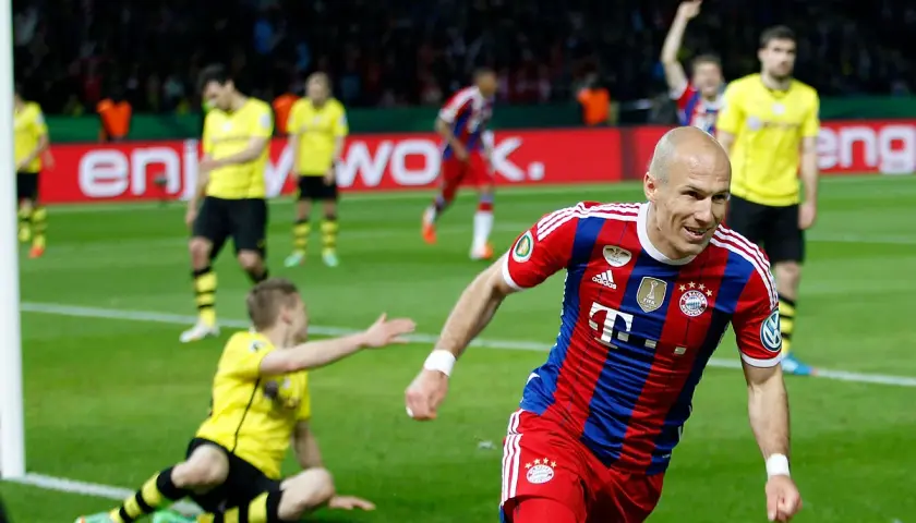Piłkarz Bayernu w czerwono-niebieskiej koszulce cieszy się z bramki. W tle zawodnicy Borussii Dortmund w żółtych strojach.