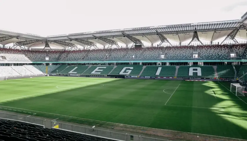 Puste trybuny stadionu Wojska Polskiego czekają na kibiców. Na zielonej murawie widać białe linie boiska.