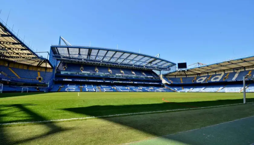 Pusta trybuna nowego stadionu Chelsea, z zieloną murawą i niebieskimi siedzeniami.