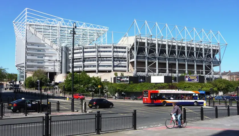 St James' Park w Newcastle, z charakterystyczną białą konstrukcją dachu, otoczony zielenią i ruchliwą ulicą z autobusem i rowerzystą.