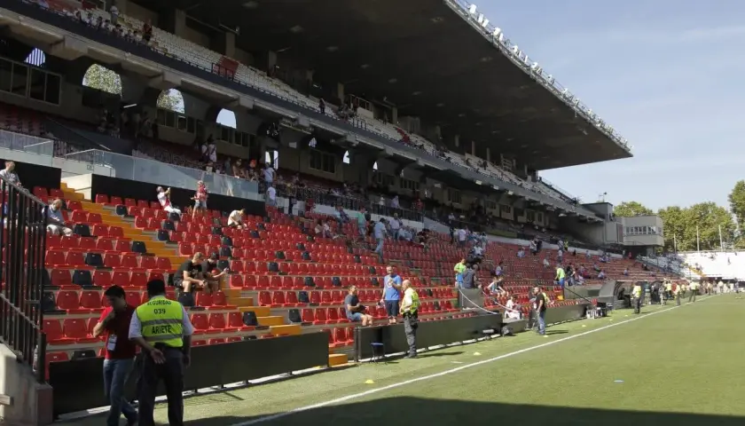 Puste trybuny stadionu Rayo Vallecano czekają na kibiców. Widać zieloną murawę i kilka osób na schodach.