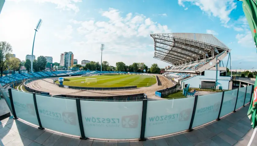 Stadion miejski w Rzeszowie z zieloną murawą, bieżnią żużlową i niebieskimi trybunami. W tle budynki mieszkalne i błękitne niebo z chmurami.