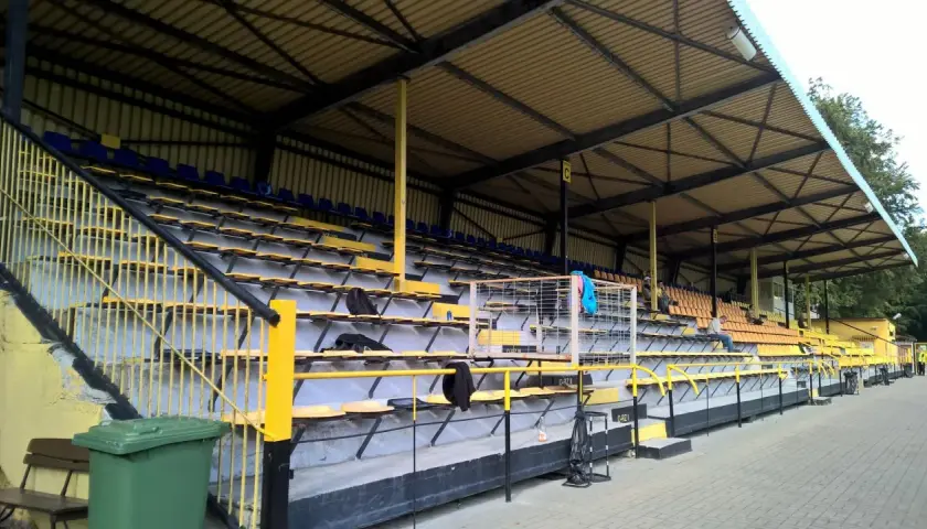 Puste trybuny stadionu Gryf Wejherowo, z żółtymi i niebieskimi siedzeniami pod zadaszeniem.