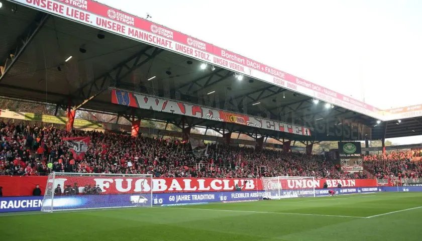 Trybuna na stadionie Union Berlin wypełniona kibicami. Na czerwonym banerze widnieje napis "UNSERE LIEBE. UNSERE MANNSCHAFT. UNSER STOLZ. UNSER VEREIN.".