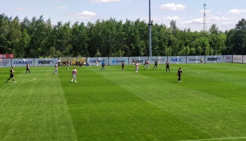 Piłkarze grają na boisku podczas meczu Betclic 3 liga. Słońce świeci, a drzewa tworzą tło.