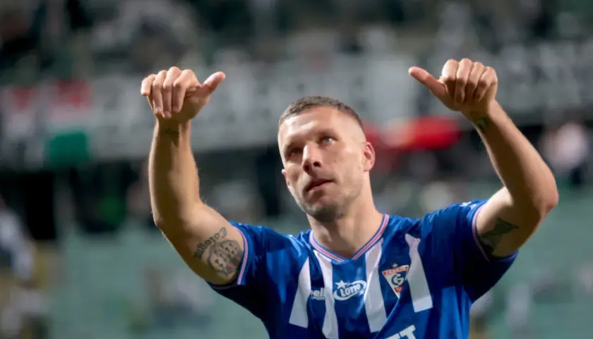Piłkarz Louis Gabriel Podolski w niebieskiej koszulce z białymi pasami, z uniesionymi kciukami, świętuje zwycięstwo na boisku.