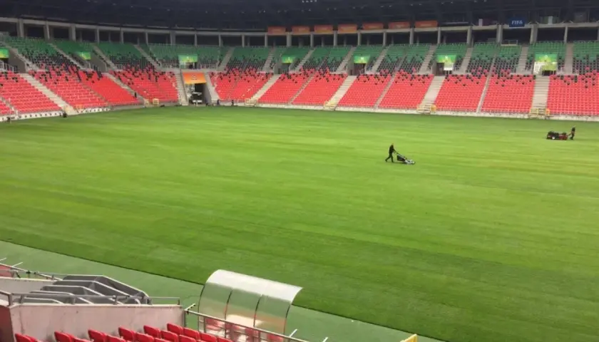 Pracownicy koszą trawę na boisku stadionu GKS Tychy. Puste trybuny w kolorach czerwonym i zielonym czekają na kibiców.