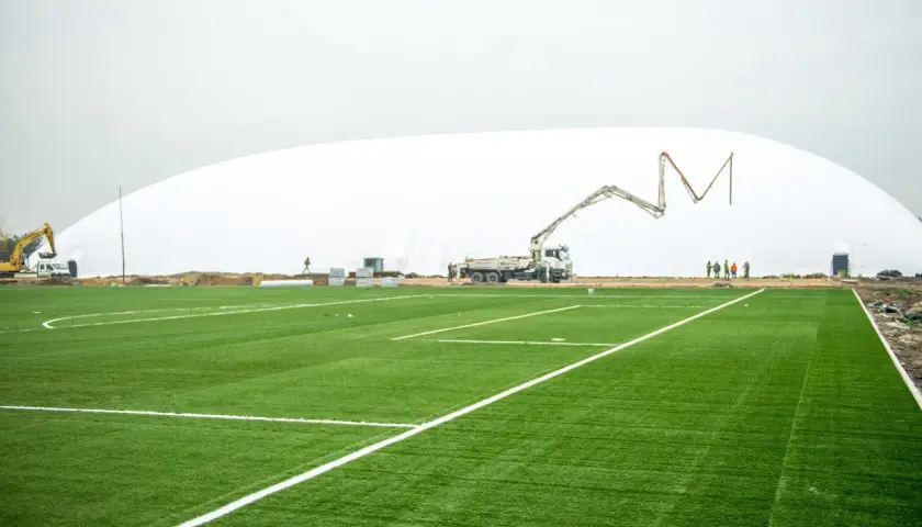 Budowa hali pneumatycznej na stadionie Ślęzy Wrocław. Sztuczna murawa z białymi liniami, koparka i gruszka do betonu w tle.