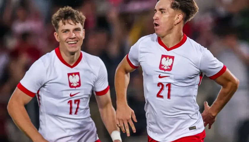 Dwóch młodych zawodników w białych koszulkach z orłem na piersi, z numerami 17 i 21, cieszy się po zdobytej bramce.