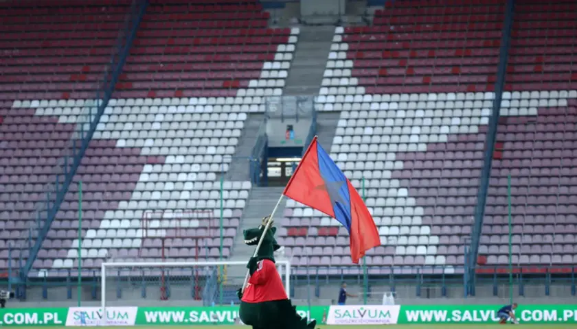 Maskotka Wisły Kraków z flagą na tle stadionu.