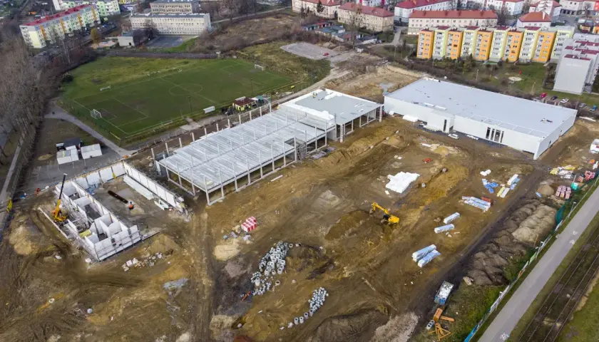 Budowa obiektu sportowego w Radzionkowie. Widać plac budowy z fundamentami, konstrukcją hali, maszynami budowlanymi i pobliski stadion.