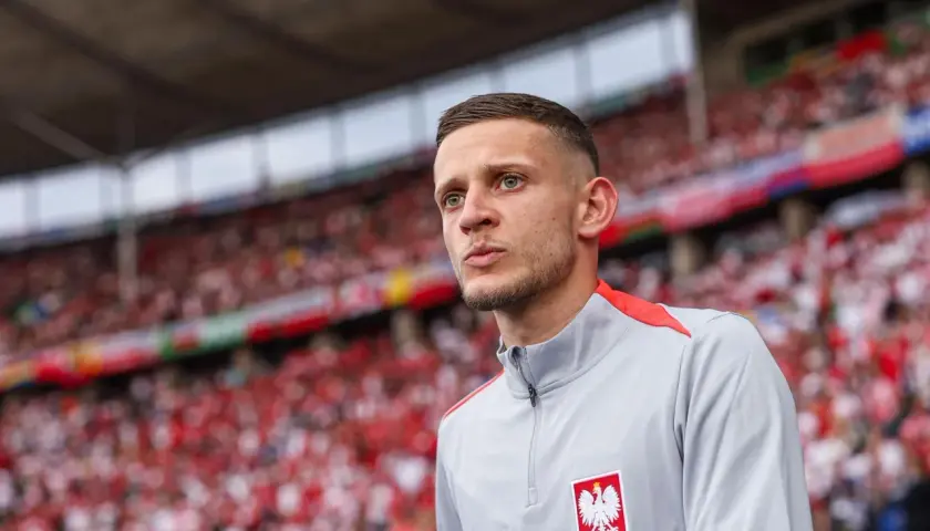Sebastian Szymański w szarej bluzie z orłem na piersi, na tle trybun wypełnionych kibicami w biało-czerwonych barwach.