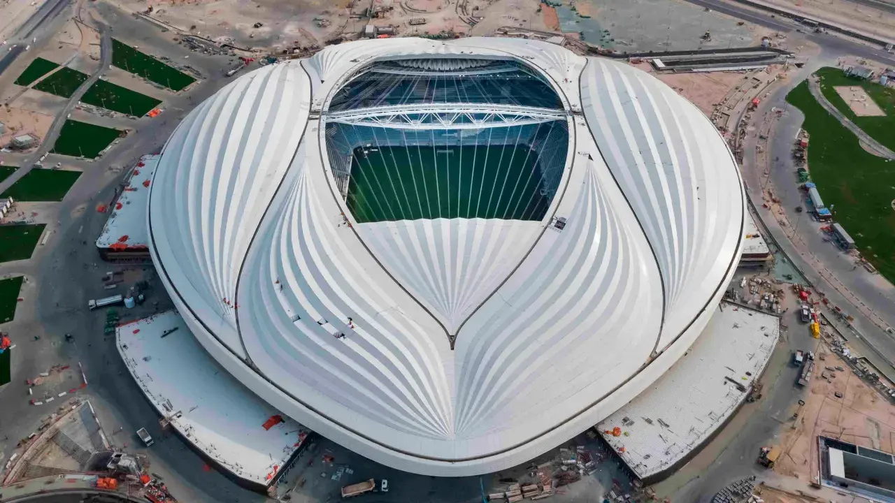 Al Janoub Stadium z lotu ptaka, z charakterystycznym białym dachem przypominającym żagiel. Widać zieloną murawę boiska i otaczające go tereny budowy.
