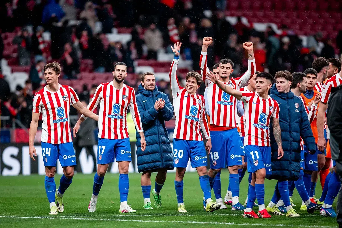 Piłkarze Atletico Madryt w pasiatych koszulkach świętują zwycięstwo. Wszyscy uśmiechnięci, niektórzy unoszą ręce w geście radości, kiedy gra Atletico Madryt przynosi sukces.