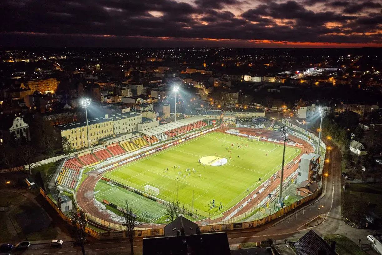Wieczorny mecz na stadionie miejskim w Chojnicach. Piłkarze biegają po jasno oświetlonej murawie, a nad miastem zapada zmrok.
