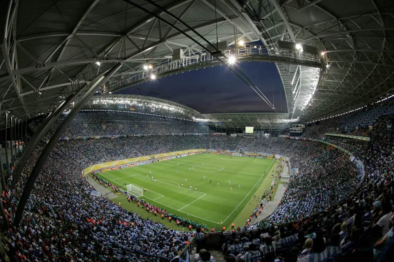 Widok z góry na pełny stadion Red Bull Arena Leipzig, gdzie tysiące kibiców w zielonych i niebieskich barwach dopingują swoje drużyny podczas nocnego meczu.