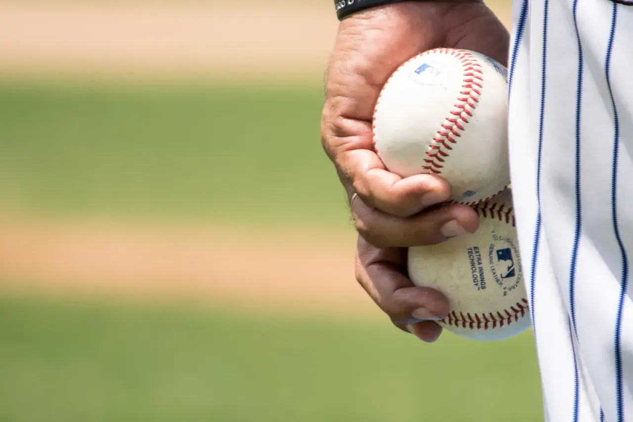 Dłoń trzyma dwie piłki baseballowe, gotowe do gry. Widać napis "EXTRA INNINGS TECHNOLOGY", świadczący o zaawansowanej technice niskiej piłki.