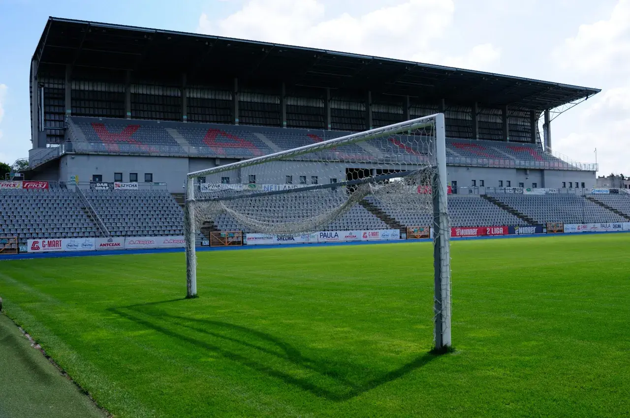 Pusta bramka na zielonej murawie stadionu KKS Kalisz. W tle trybuny i zadaszenie.