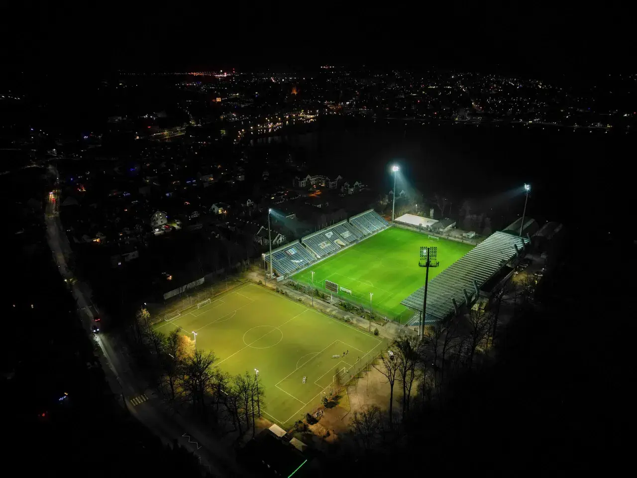 Nocne oświetlenie rozświetla zieloną murawę stadionu Sokoła w Ostródzie, otoczonego ciemnością i miejskimi światłami.