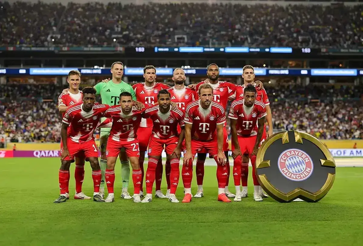 Drużyna Bayern Monachium – zawodnicy w czerwonych strojach pozują do zdjęcia na tle trybun. Obok nich stoi okrągłe logo klubu.